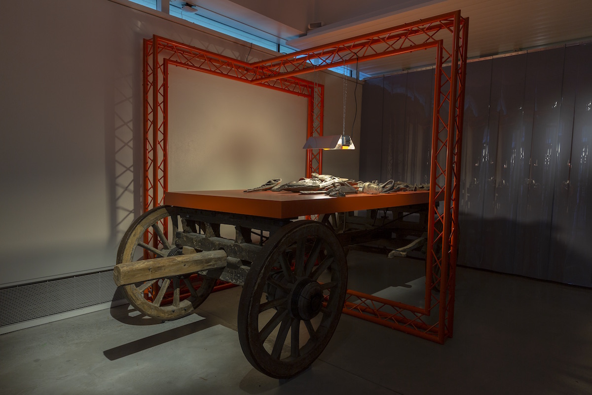 Large orange truss structure on top of a wooden timber carriage. There is a light hanging down highlighting a pile of objects on an orange table
