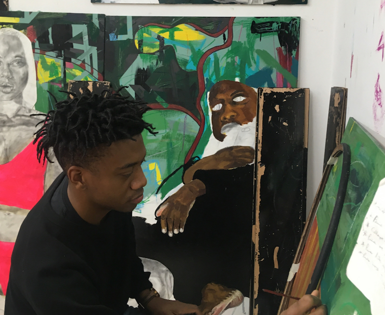Jerome. crouched down and painting with another of his artworks in the background