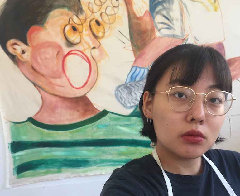 Sae Yeoun Hwang looking out at the viewer with one of her artworks in the background
