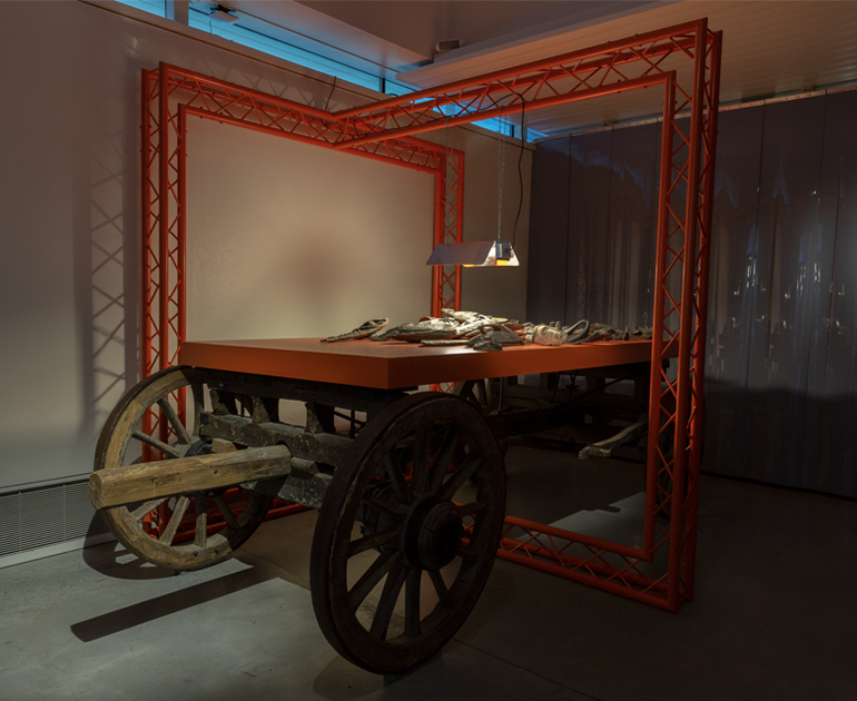 Large orange truss structure on top of a wooden timber carriage. There is a light hanging down highlighting a pile of objects on an orange table