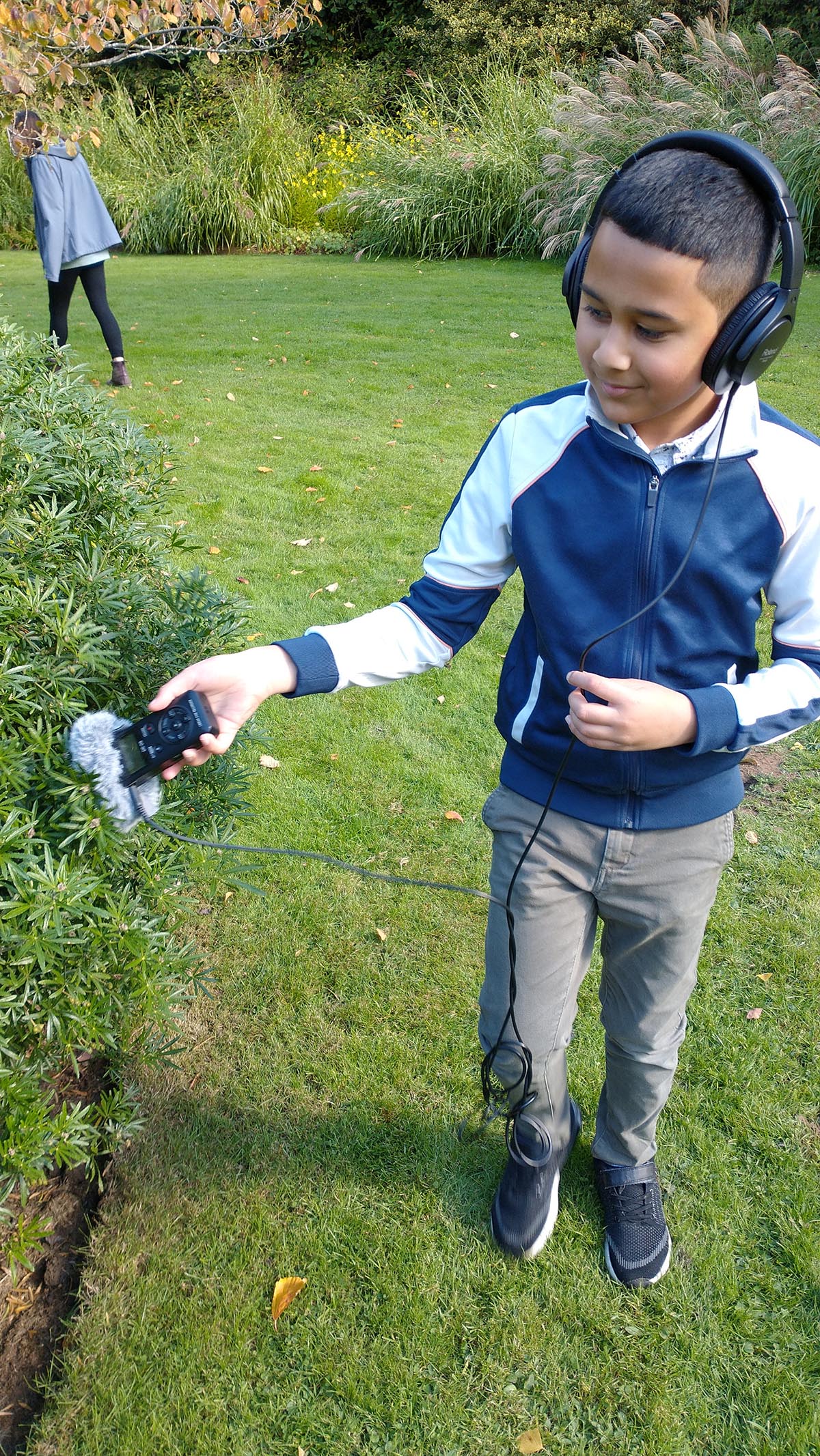 Boy, wearing headphones and a white and blue coat, holding a piece of audio equipment towards a plant