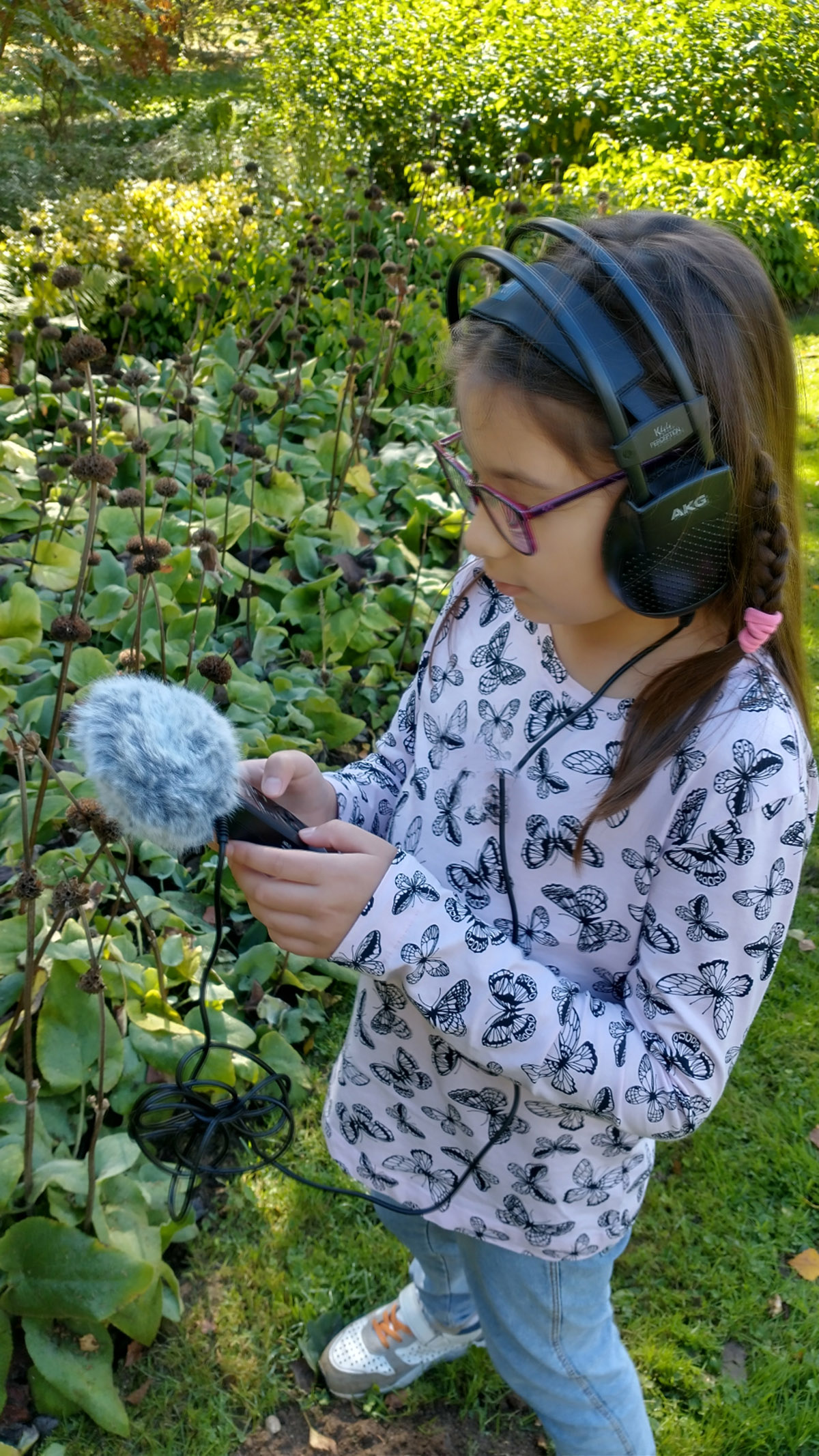 Girl, wearing headphones and a white top with blue butterflies on it, holding a piece of audio equipment towards a plant