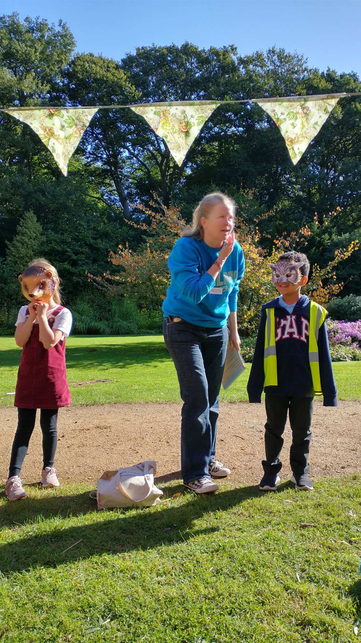 Two children, wearing animal face masks, and a blonde woman wearing a blue jumper, standing underneath bunting.