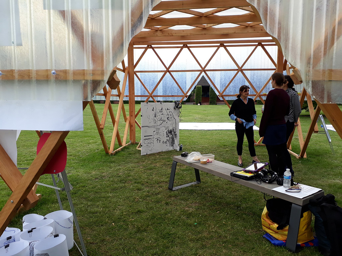 Several people inside the Urban Room, a wooden structure with plastic sheeting on the top half. The people are surrounded by a variety of art and standing next to a grey bench with paper, a bottle and some digital equipment on it.