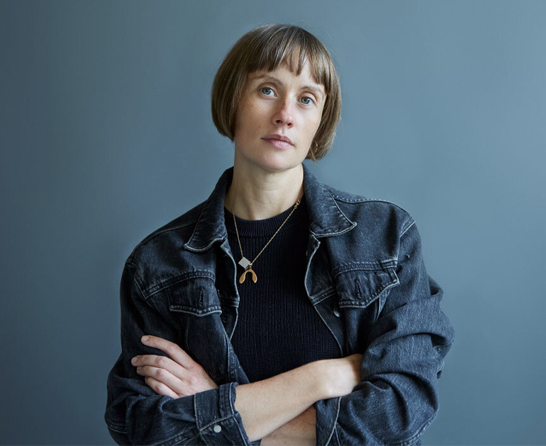 A woman with short light brown hair, wearing a denim jacket, with her arms crossed.