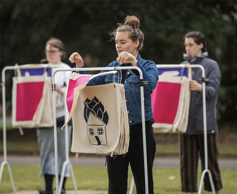 3 figures with metal structures with fabric squares in the centre that appear to work like a flip chart. The figures in the centre is currently 'flipping' a square with a burning building visible on the square underneath