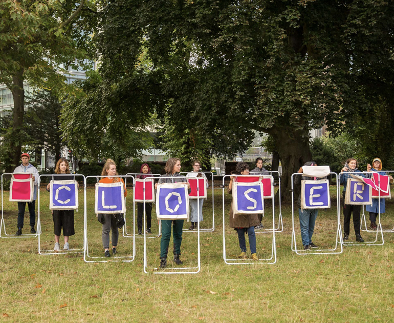 11 figures with metal structures with fabric squares in the centre that appear to work like a flip chart. The figures at the front have spelled out the word Closer using their fabric squares.