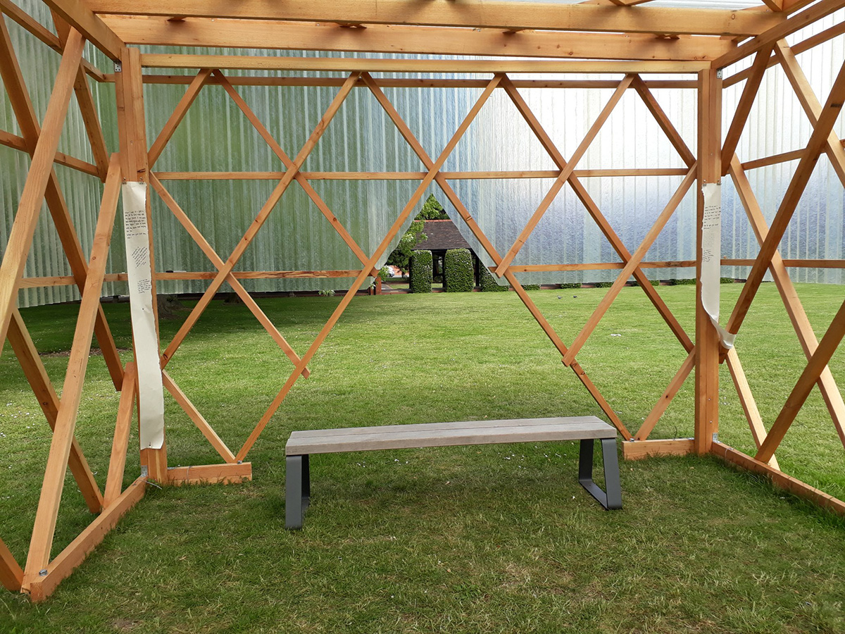 Inside of the wooden Urban Room structure with plastic on the top half. There is an empty grey bench by a triangular opening with long pieces of paper attached to the structure on either side