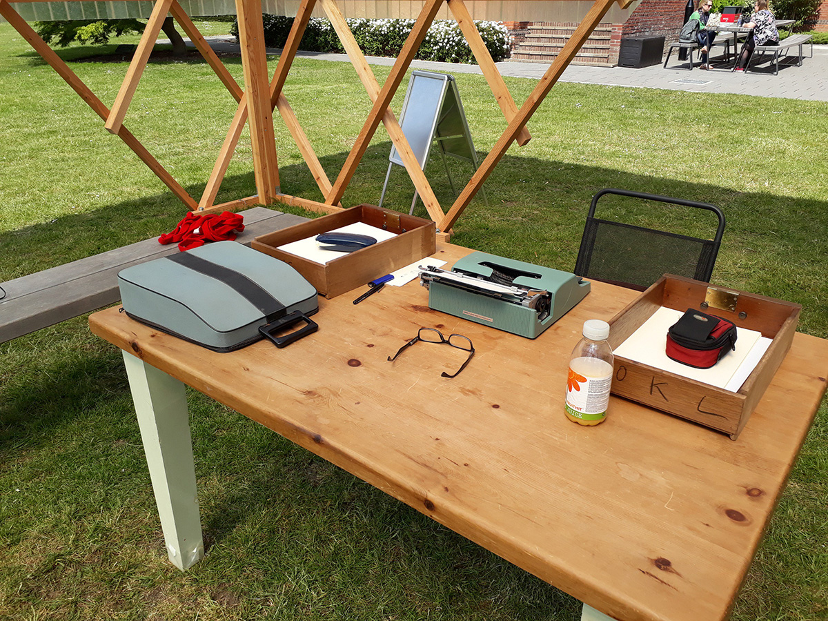 Wooden table with a typewriter, case, two wooden boxes of paper, a pair of glasses and a bottle. Next to the table is a black chair and a grey bench