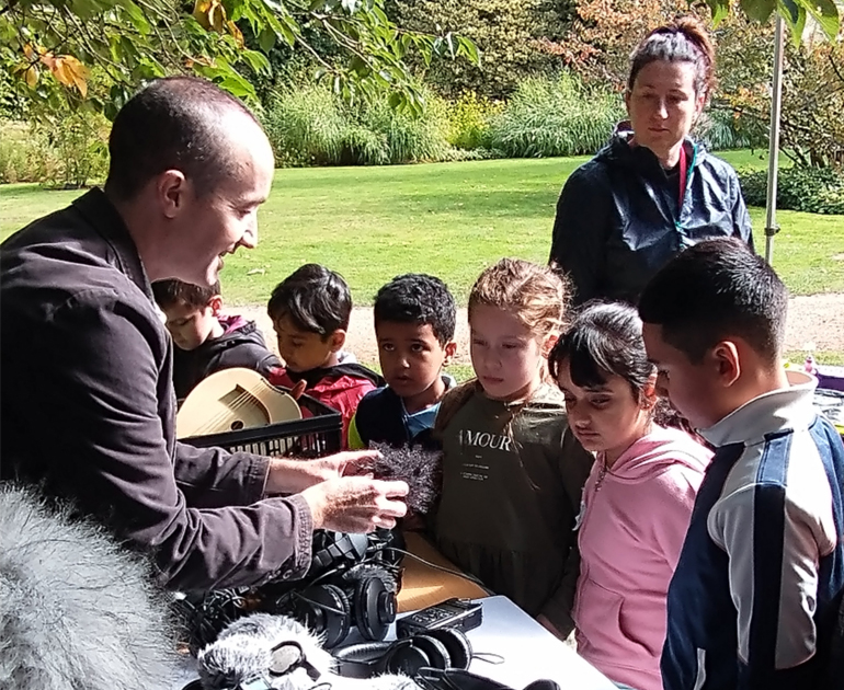 Six children standing at a table looking at instruments and sound equipment. There is a man showing the equipment to the children and a woman standing behind the children observing