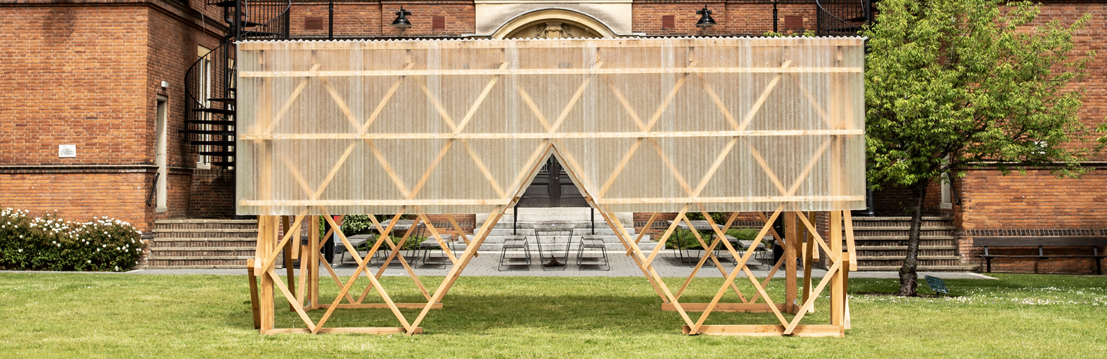 Exterior view of the Urban Room, a wooden structure which is open at the bottom and has plastic sheeting wrapped around the top half. There are large triangular openings at the bottom to access the inside of the structure