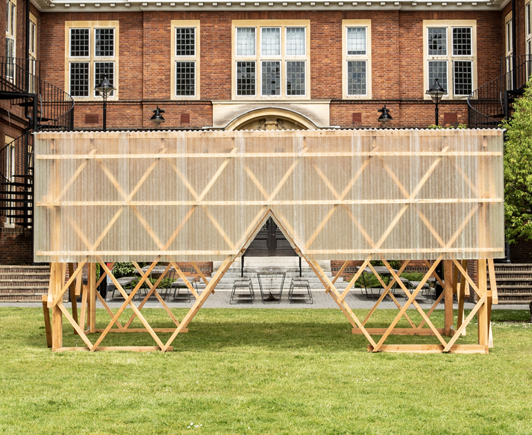 Exterior view of the Urban Room, a wooden structure which is open at the bottom and has plastic sheeting wrapped around the top half. There are large triangular openings at the bottom to access the inside of the structure