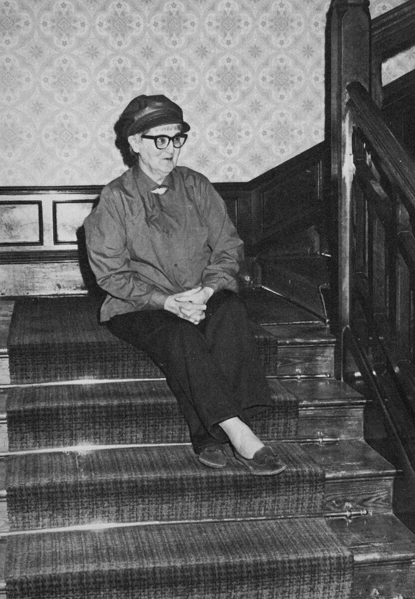 Kathleen Hale sitting on the top of a set of stairs. She is wearing a top, trousers, hat and glasses. On her right a second set of stairs continues upwards.
