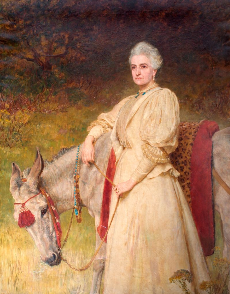 Almost full-length, life-size image of elderly Lady Wantage, in a yellow dress, standing by grey donkey with red and leopard-skin trappings, in a rural setting.