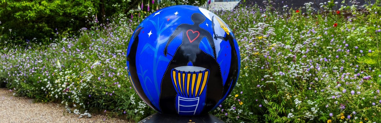 Painted globe, with several figures. One is a solid black figure with a outlined heart standing behind a drum. To the right is a figure that has a detailed face and is holding a sword. The globe is outside in front of a bed of wild flowers.