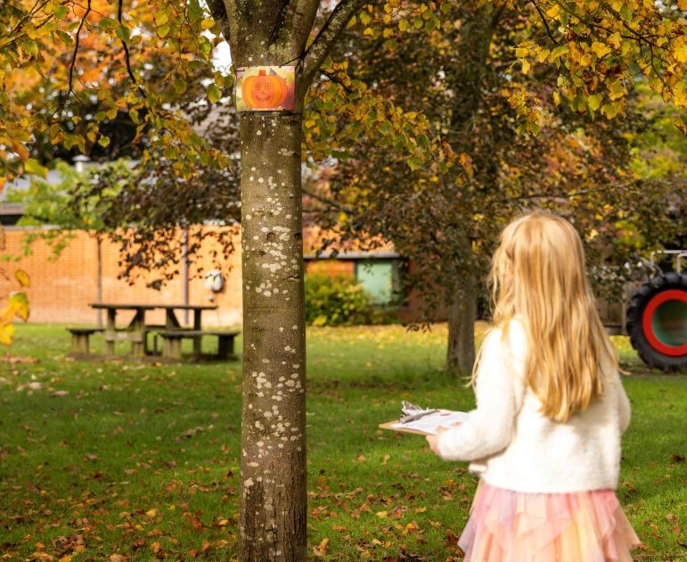 Back of a standing child holding a clipboard. The child is outside next to a tree. There is a picnic table in the background.
