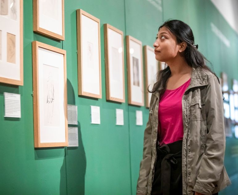 Woman looking at a series of drawings on display in an exhibition. The wall behind is painted green.