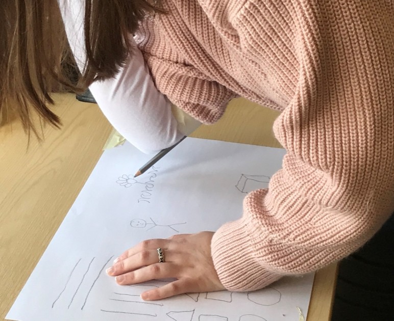 A woman leaning over a piece of paper. There is a pencil attached to her right elbow. She is using this pencil to draw.