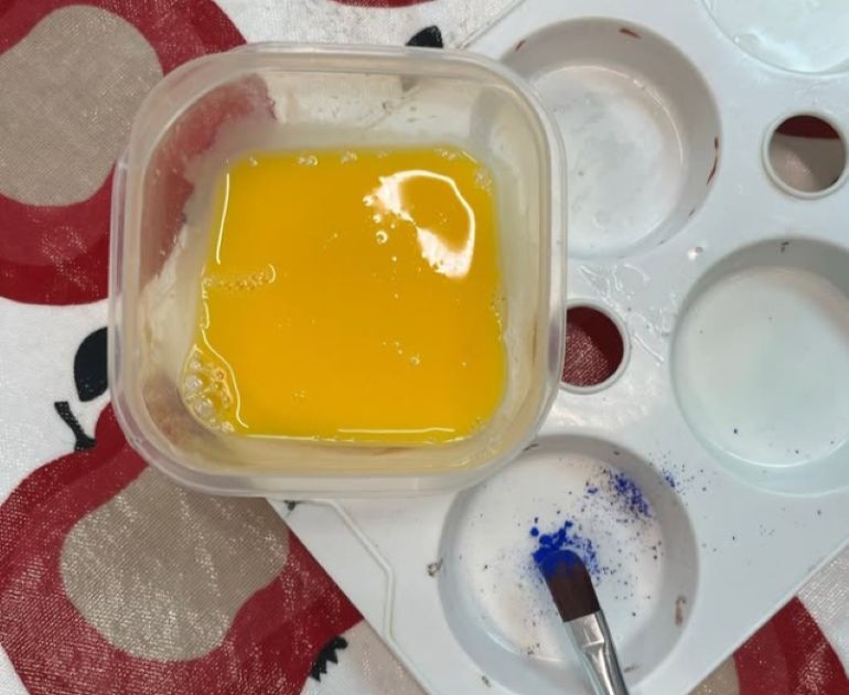 Egg yolk in a plastic tub on top of a plastic painting palette. There is a paint brush in the bottom with blue pigment on it.