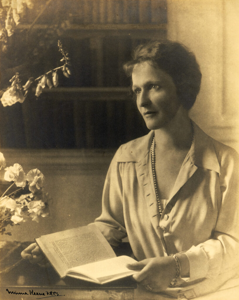 A sepia portrait by photographer, Minna Keene, showing Nancy Astor seated at a table with a book.