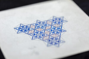 A close-up of a geometric drawing on paper featuring an intricate Islamic-style star pattern. The design is composed of interlocking blue and red triangles and hexagons, forming repeating six-pointed stars and floral motifs.