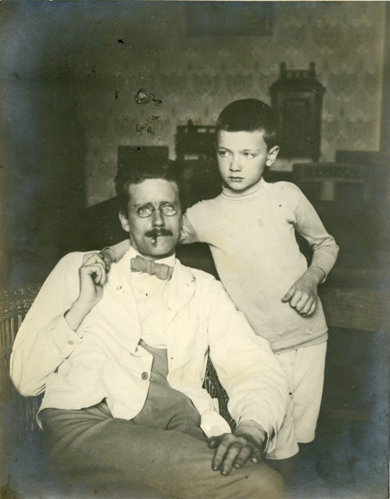 James Joyce is sitting on a chair. His young son has his arm around his father's neck and they are holding hands.