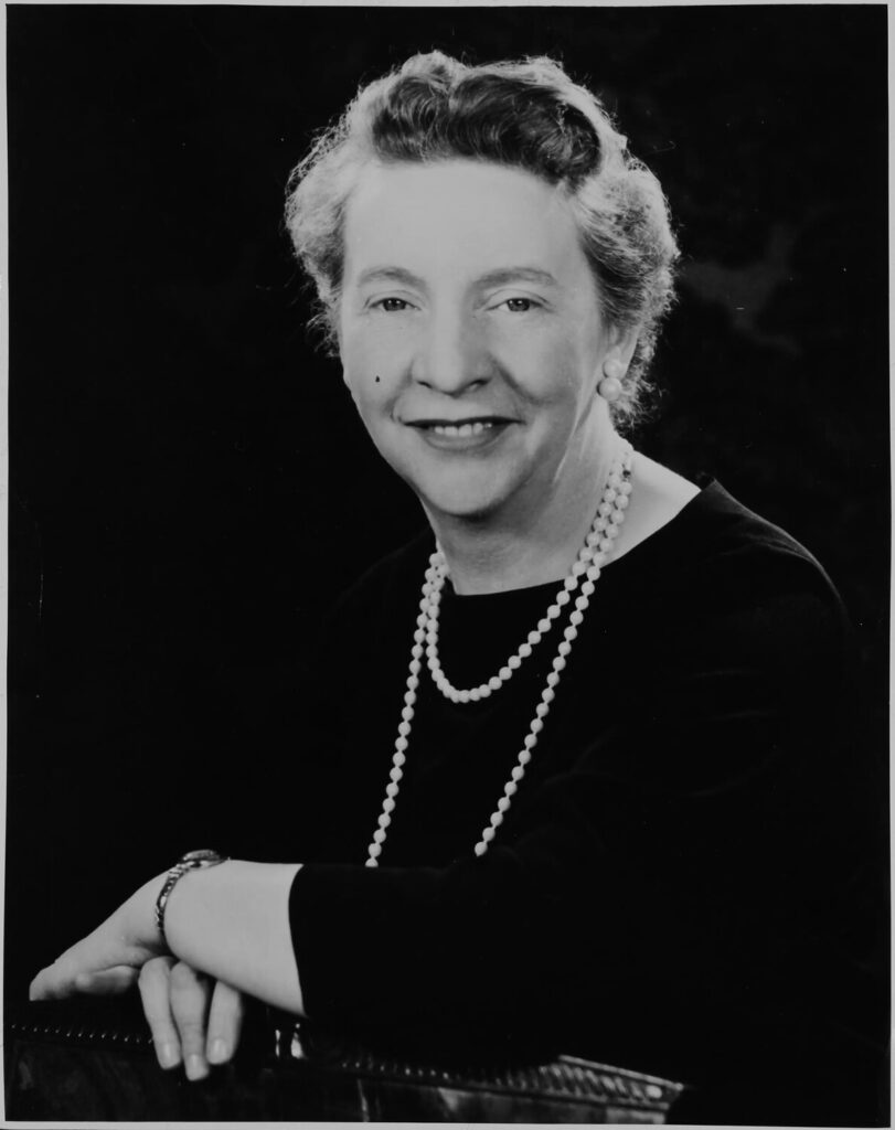 Black-and-white studio portrait of a woman seated with her arms crossed, wearing a dark dress and a long pearl necklace