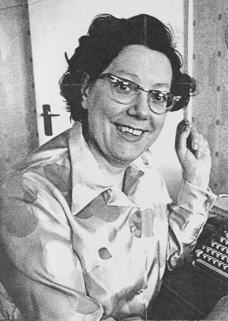 Black-and-white portrait of a woman wearing glasses, seated at a typewriter and smiling at the camera