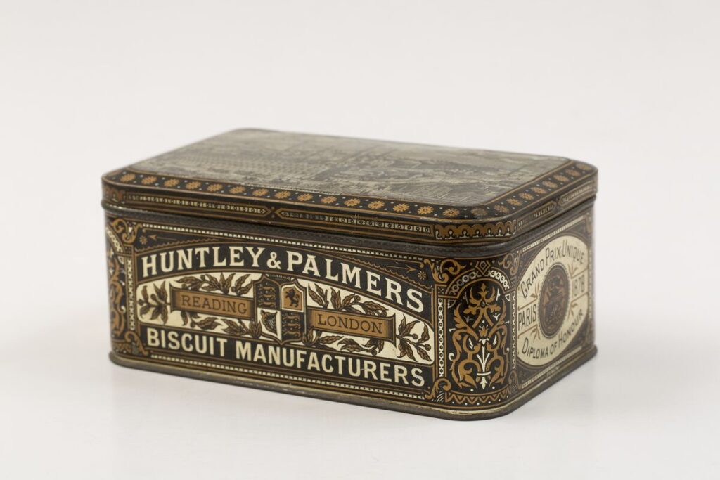 Rectangular decorative biscuit tin made of metal, shown on a plain light background. The tin is richly patterned in brown, gold, and cream, with ornate floral and scroll designs. One side clearly displays the text “Huntley & Palmers, Biscuit Manufacturers” with banners reading “Reading” and “London.” The lid is slightly domed and decorated with intricate printed imagery.