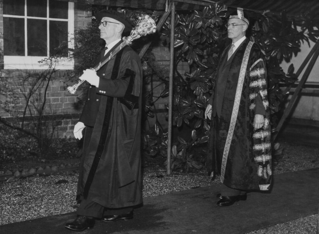 Black-and-white photograph showing two people in formal academic robes walking outdoors along a paved path. The person in the foreground carries an ornate ceremonial mace, while the person behind wears a long robe with a patterned stole.
