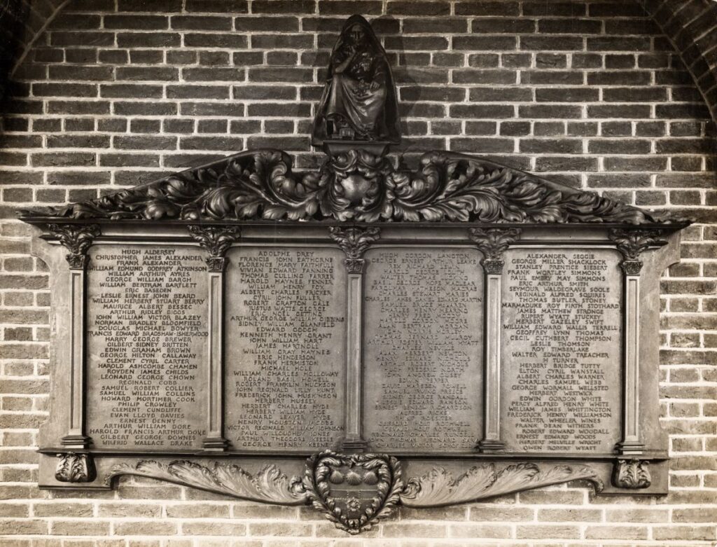 Large carved memorial plaque mounted on a brick wall. The memorial features 4 panels engraved with names in capital letters, with the University coat of arms at bottom centre and a figure of veiled seated woman in attitude of sorrow at the top centre.
