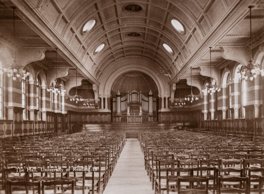 Sepia-toned photograph of a large, ornate hall with rows of wooden chairs facing a pipe organ.