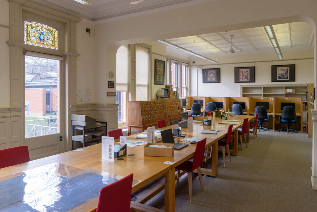 The reading room at The Museum of English Rural Life, and University of Reading, Special Collections; wooden research tables with red chairs, individual computer workstations, a card catalogue cabinet, and a stained glass peacock window.