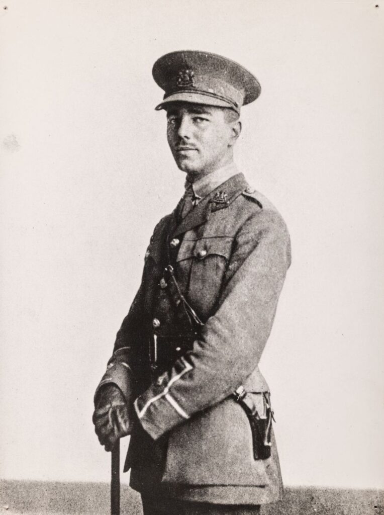 Black and white photograph of Wilfred Owen in First World War army uniform, standing three-quarter length and turned slightly to the side.
