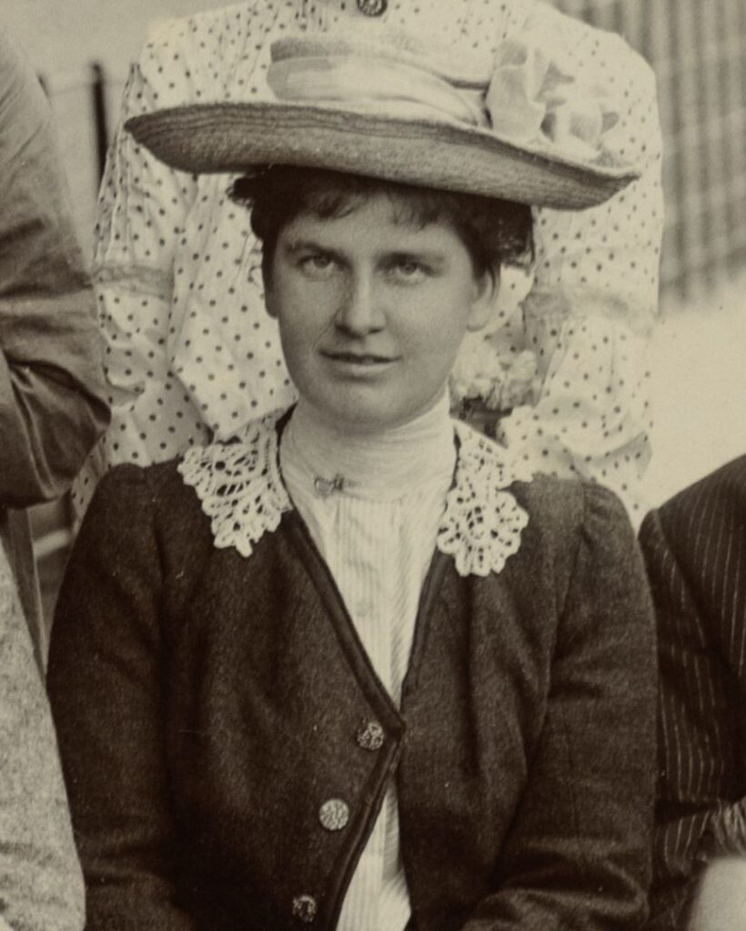 Black and white close-up photograph of Mabel Rayner, cropped from a group portrait, wearing a wide-brimmed hat with decorative trim, a lace collar, and a dark buttoned jacket.