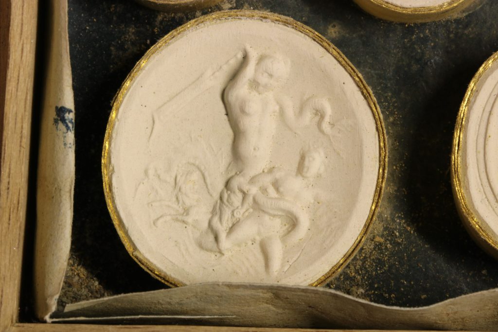 The gem cast has a raised surface that shows Scylla attacking a sailor. The sailor's body is facing the right and his head is turned back, looking at Scylla. The sea monster stands over him with her right arm raised in a threatening stance.