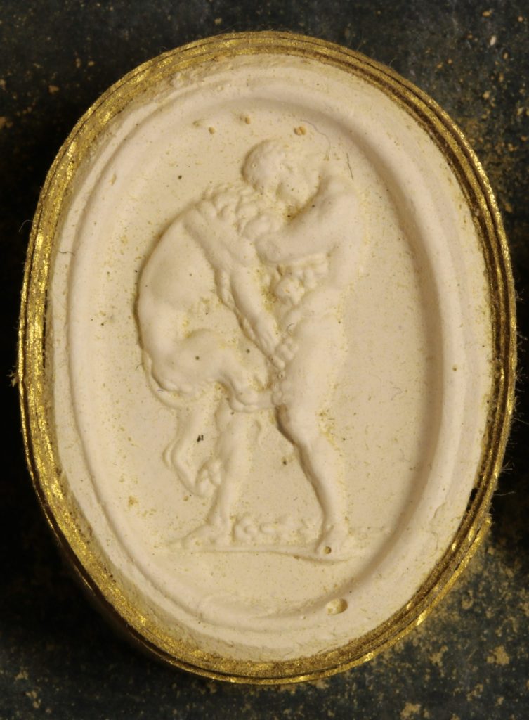 The gem cast has a raised surface that shows Hercules fighting the Nemean Lion. Hercules is the figure on the right, who is holding the Nemean Lion off of the ground. The lion is trying to free itself by using its forearms to push away from Hercules.