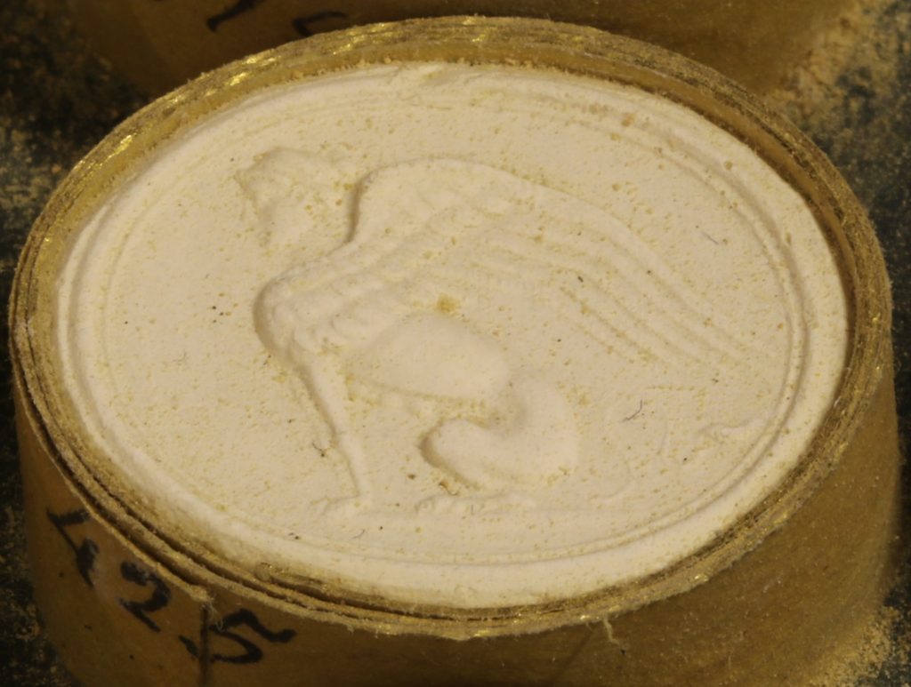 A gem cast that has a raised surface. Shows a side profile of a seated sphinx that is facing the left.