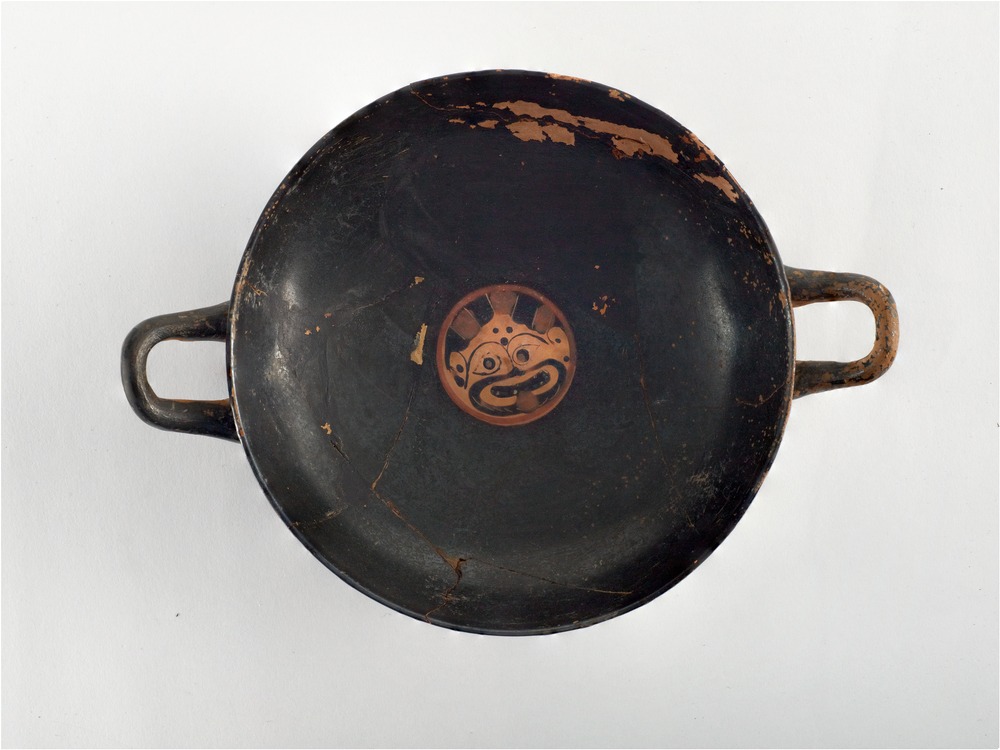 Image shows a round black-figure cup that has two handles. In the centre of the cup, an image of Medusa's face is visible and she has her tongue out. The rest of the cup is black, with the exception of a slight wear.