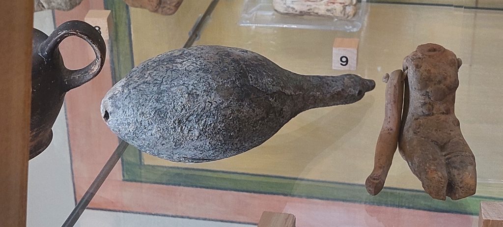 A glass shelf in a museum with a clay rattle on it. The rattle is grey in colour and made of ceramic. It is shaped like a gourd, with a long handle or neck. 